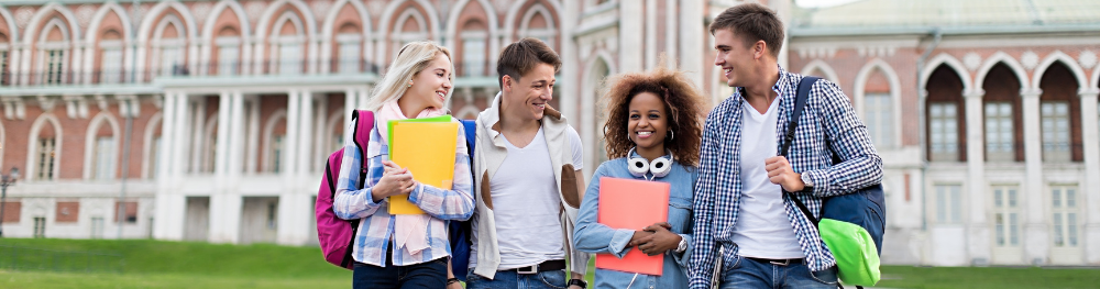 Four students with books and backpacks standing in front of a university building.